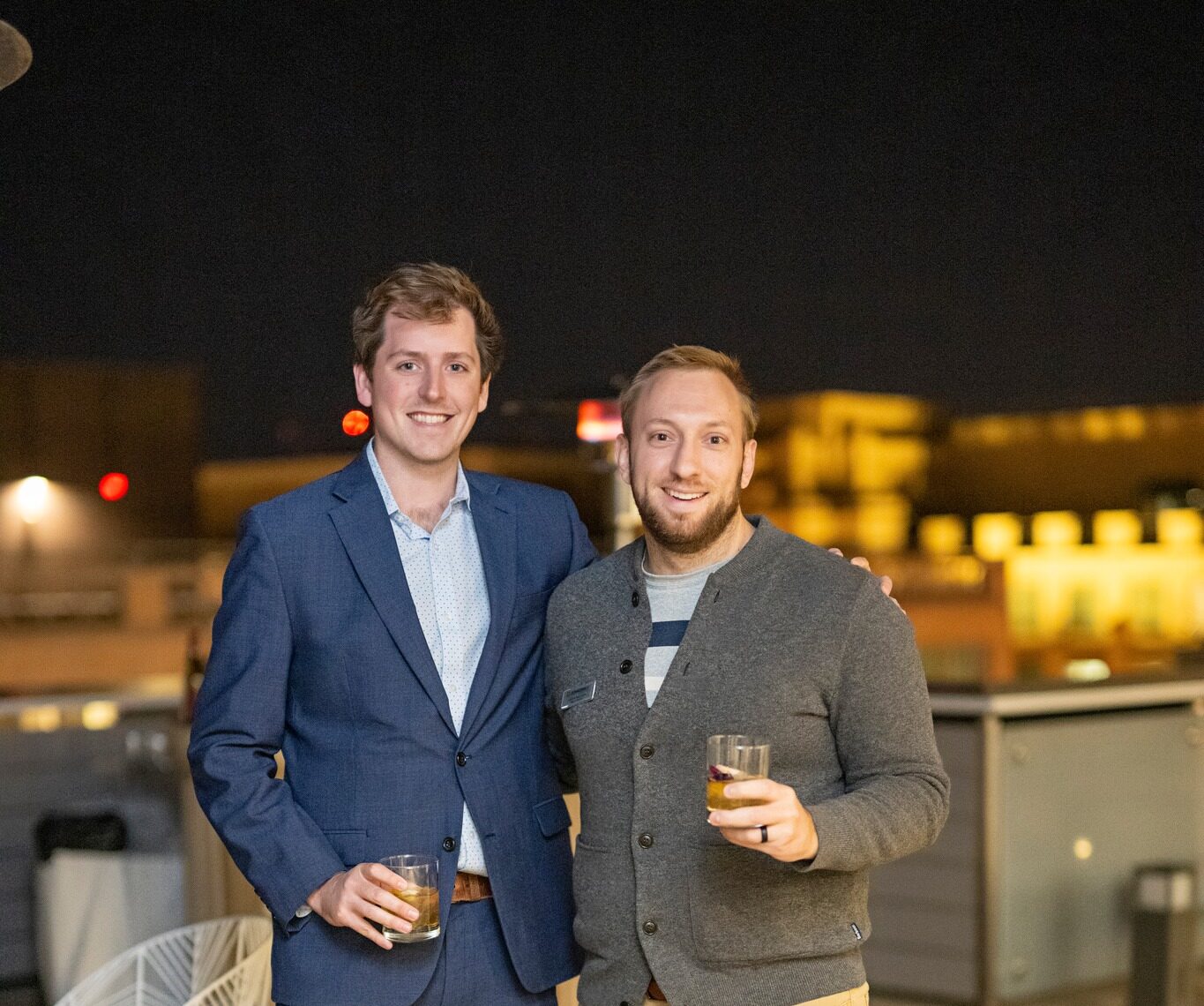 Two men at a rooftop cocktail party with a view of city behind them