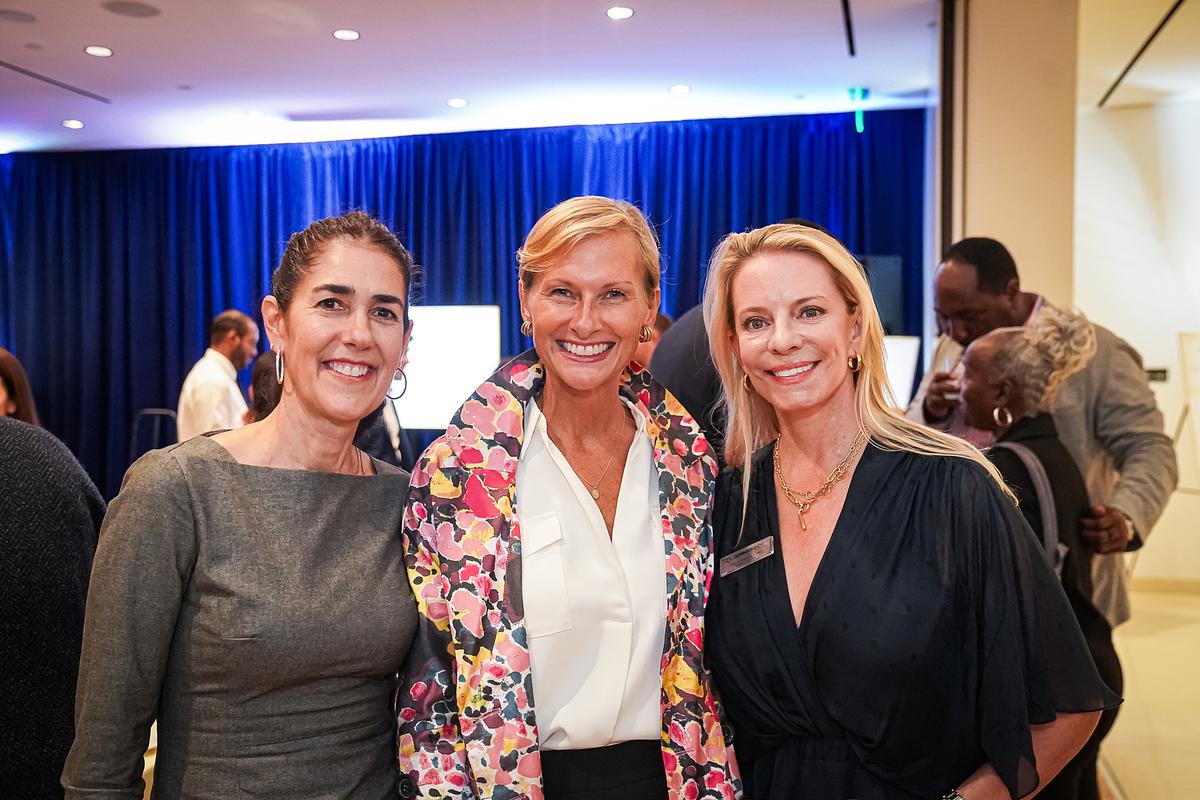 Three women pose in front of blue curtains at a DowntownDC event, with others mingling behind them